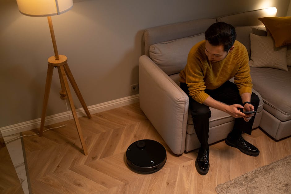 Man using smartphone in a cozy living room with smart robotic vacuum and warm lighting.