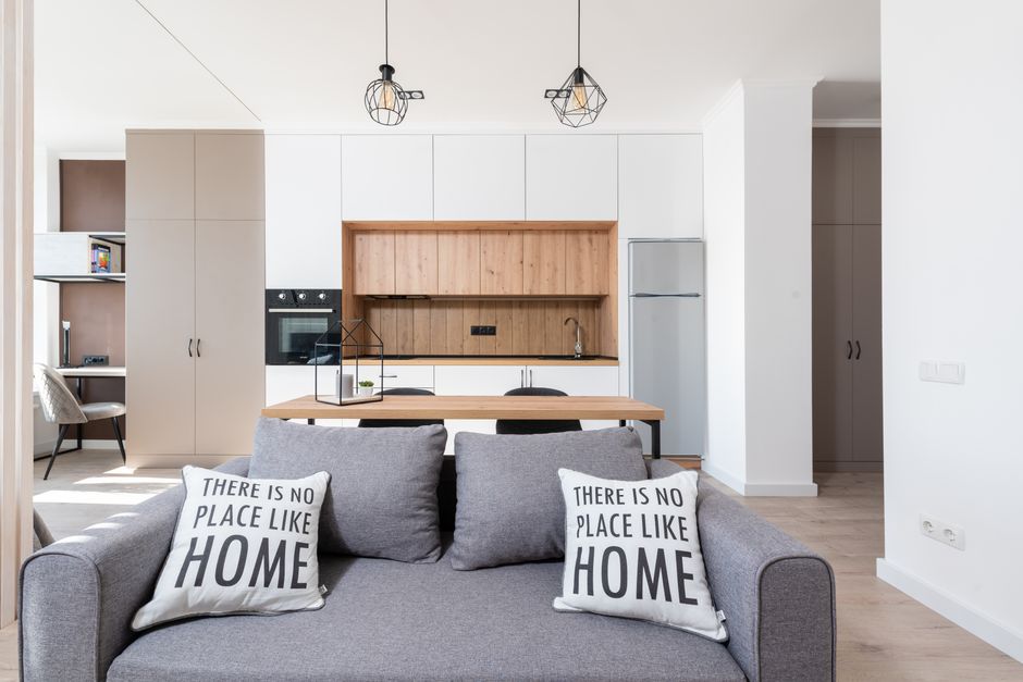 House interior with couch with pillows near table and open kitchen with cabinets and oven with fridge