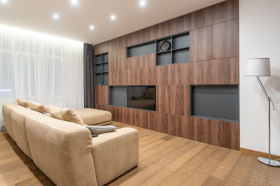 Comfortable sofa with cushions placed on parquet near cupboards with decorative black shelves in spacious living room with tulle on window